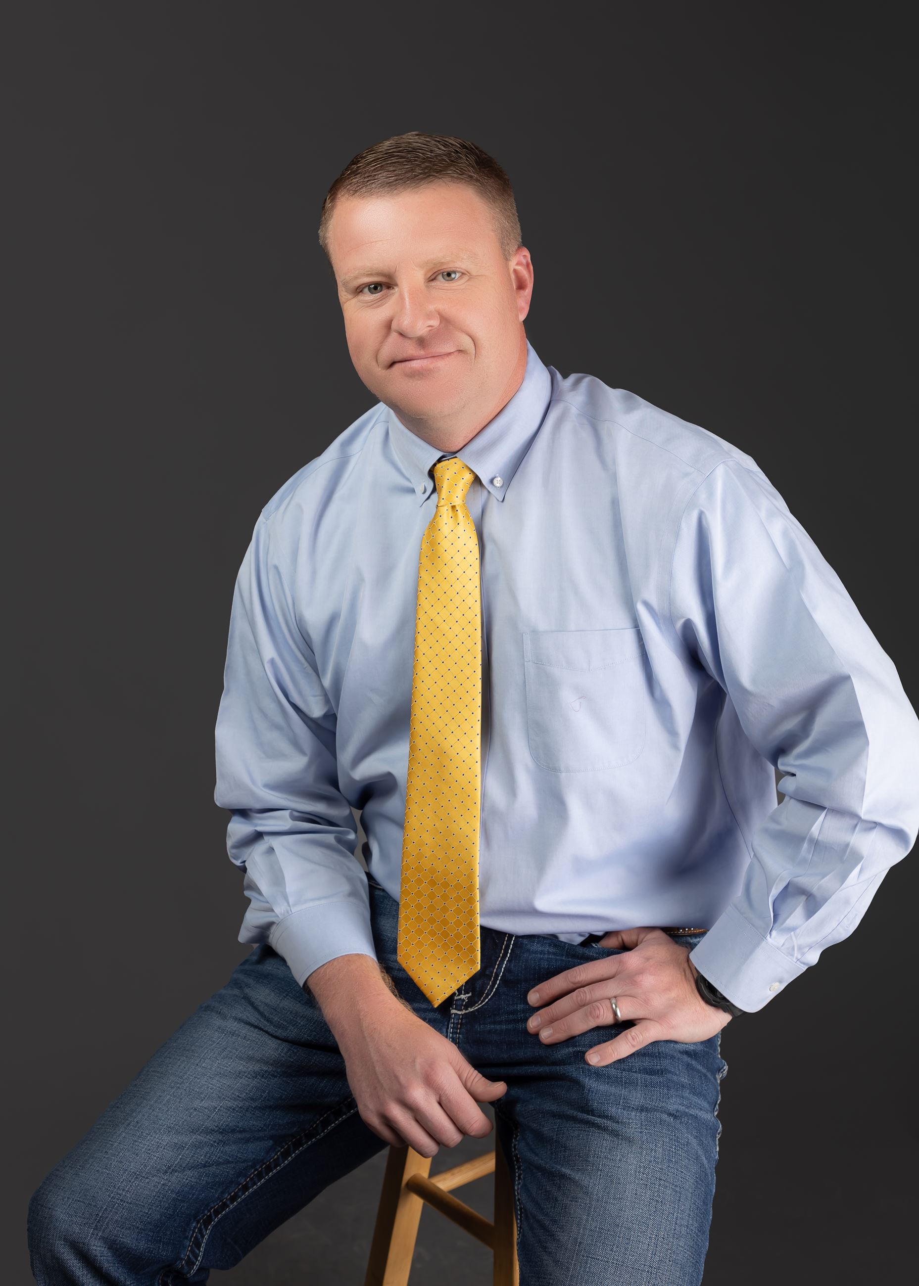 Mayor Jon Henry pictured sitting on stool in light blue button up and yellow tie